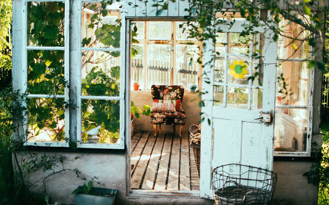 A cozy, sunlit greenhouse with large glass windows and an open door. Inside, there is a floral-patterned armchair with colorful cushions, wooden floorboards, potted plants on the windowsill, and a basket on the floor. Greenery grows around and inside the greenhouse, creating a peaceful, inviting atmosphere.