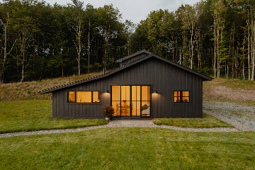 A modern, single-story dark wood house with large windows and warm interior lighting, situated on a grassy plot with a gravel pathway and surrounded by dense forest.