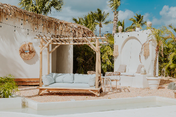 A stylish outdoor lounging area featuring a light wooden pergola with a cushioned sofa underneath, tropical palm trees in the background, a thatched roof, and decorative elements such as hanging lights and straw wall art. There are also white walls, patio furniture, and a shallow water feature in the foreground, all set under a partly cloudy sky.